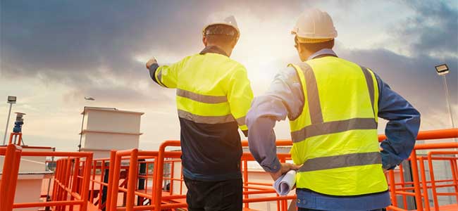 two people in yellow safety vets and a white hard hat stand near orange barricades with a building in the background. The sky, where one of the people is pointing to, is grey, but the sun appears between the people